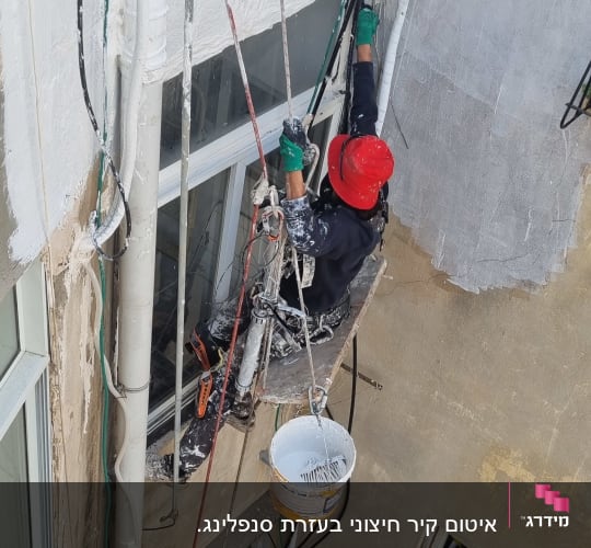 עובד בגובה עם קסדה אדומה מבצע עבודת צביעה על בניין